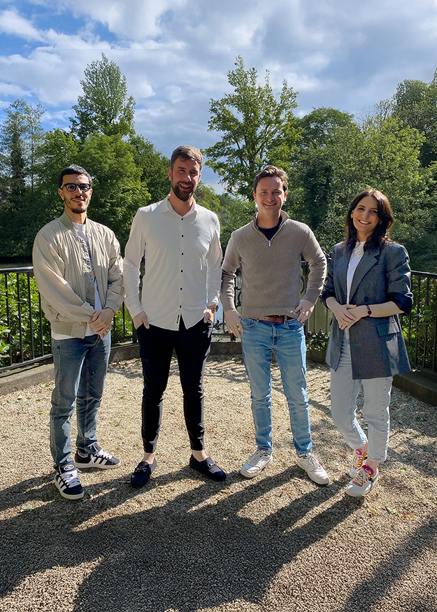 Team isales Vier collega's van ZorgDomein staan buiten in de zon, lachend en ontspannen, met een groene, bosrijke achtergrond.